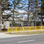 Zdjęcie przedstawia budynek Szkoły w Chotyczach od strony jezdni. Wokół chodnika znajdują się czerwone i żółte barierki bezpieczeństwa. Przed budynkiem rosną wysokie sosny.