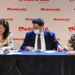 Zdjęcie przedstawia trzy osoby składające podpisy na umowach. Siedzą przy stole nakrytym czarnym obrusem. Pierwsza od lewej to Ewa Janina Orzełowska, obok niej Burmistrz Mariusz Kucewicz oraz Skarbnik Marta Stasiuk.